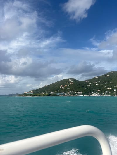 The image presents a wide-angle view of Road Town, Tortola, British Virgin Islands, observed from a low vantage point on a moving body of water. In the immediate foreground, a segment of a white, curved tubular railing is visible on the lower right, with white aerated water and spray indicating motion. The foreground and midground consist of a vast expanse of clear, turquoise-blue ocean water, exhibiting minor surface ripples. The background is defined by a continuous chain of verdant, densely vegetated hills rising directly from the water's edge, extending across the entire horizon. Numerous light-colored residential and commercial structures are sparsely scattered across the slopes and bases of these hills. In the central-right portion of the background, a concentration of white-hulled sailing vessels and yachts is moored within a visible marina or harbor area. The sky occupies the upper portion of the frame, featuring a mosaic of bright blue patches interspersed with various white and grey cloud formations, including wispy cirrus and denser cumulus clouds, particularly prevalent over the left and central landmasses. No identifiable individuals or explicit interactions are discernible within the frame.