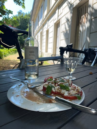 An outdoor table setting is depicted in this scene, featuring a partially eaten caprese salad on a white plate, a fork, and a knife. A glass of white wine sits next to a bottle of white wine labeled "BANDOL 2023" from "Domaine de la Tour du Bon." In the background, a bicycle is parked, with a building visible behind it. The setting appears to be in Bièvres, France, given the context of the wine and the architectural style of the building. The overall atmosphere suggests a leisurely meal enjoyed outdoors.