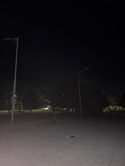 An outdoor, wide-angle, eye-level shot captures a dimly lit park scene at night in Egorlykskaya, Russia. The foreground is dominated by a paved walkway with a repeating geometric pattern of light-colored bricks. A circular manhole cover is visible on the walkway in the lower right quadrant of the image. To the left of the walkway, a tall, slender street lamp with a white pole and a black light fixture stands prominently. Beyond the walkway and street lamp, a grassy area transitions into a dense cluster of trees, their foliage dark and indistinct against the night sky. Another street lamp, similar in design to the first, is visible further back in the middle ground, to the right of the first. Its light casts a faint glow on the surrounding trees and ground. In the background, beyond the trees, there are suggestions of structures or distant lights, too indistinct to identify. The overall atmosphere is serene and still, with no people or animals present in the frame. The sky is a deep, starless black.