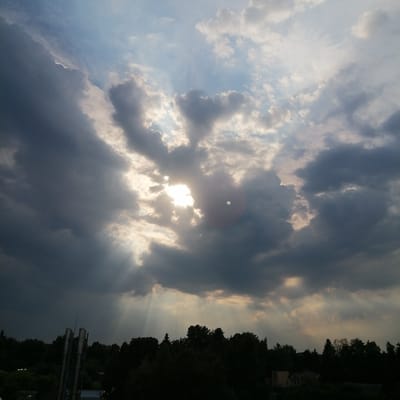 The image captures a dramatic sky over Trubacheyevka, Russia, with the sun partially obscured by dark, heavy clouds. Rays of light emanate from behind the clouds, creating a crepuscular effect. In the foreground, silhouetted against the sky, are the dark shapes of trees and some industrial or urban structures, including what appears to be a set of tall industrial chimneys on the left. The overall mood is somber, with a sense of impending weather change.
