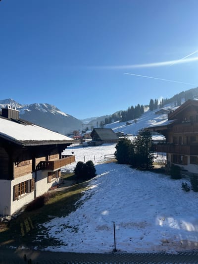 The image depicts a winter landscape in Saanen, Switzerland, under a clear, bright blue sky with a distinct airplane contrail visible in the upper right quadrant. Strong sunlight illuminates the scene from the right, casting long shadows.
In the immediate foreground, a dark, vertical pole stands in partially melted, uneven snow, which gives way to patches of green grass and brown earth. A dark, gridded surface, possibly pavement or a balcony floor, is at the bottom edge.
The mid-ground features a snow-covered field interspersed with several coniferous trees. Two prominent traditional chalet-style buildings flank this area. On the left, a multi-story building with dark wooden siding, white lower walls, multiple windows with closed brown shutters, and a wooden balcony on the second floor presents a snow-covered roof. On the right, another similar, multi-level chalet with dark wood and balconies is partially visible, also featuring a snow-laden roof. Further back in the mid-ground, a large, dark-roofed wooden barn or agricultural building is discernible in the snowy field, along with a wooden fence line.
The background consists of majestic snow-capped mountains and forested slopes. Pine or fir trees, some dusted with snow, cover the hillsides, where sections of groomed snow suggest ski runs or winter trails. Distant structures, likely houses or cabins, are scattered across these slopes. No individuals or direct human actions are observable within the frame.