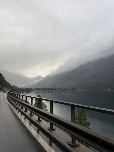 The image presents a majestic and moody landscape in Le Prese, Switzerland, viewed from a road, likely from a moving vehicle. The scene is dominated by a vast lake surrounded by towering mountains under an overcast sky.

In the foreground, a multi-layered metal guardrail runs along the right side of a wet, dark asphalt road. The road surface glistens, indicating recent rain or mist. Beyond the guardrail, a calm body of water, a lake, reflects the muted light from the sky and the dark shapes of the mountains and trees lining its shores. A solitary green-leafed tree is partially visible behind the guardrail on the right, with its reflection visible in the lake.

The midground features steep, dark mountains rising from the lake. Their lower slopes are covered in dark green foliage, which gradually gives way to rugged, rocky terrain at higher elevations. Some distant mountain peaks are partially obscured by thick, grey clouds or mist, adding to the dramatic atmosphere. Faint patches of snow can be discerned on the upper slopes of some mountains. Along the far shore of the lake, a subtle line of small structures, possibly buildings or a village, can be seen.

The sky is entirely overcast, filled with heavy, uniform grey clouds that blend seamlessly into the mountain forms in the distance. The diffused light suggests a daytime scene, though the heavy cloud cover gives it a somber, atmospheric quality. Far down the wet road, a small vehicle with visible red brake lights appears in the distance. There is no visible text in the image.