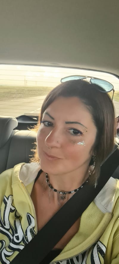 A woman with glittery makeup and a nose ring is photographed indoors, likely in a car given the visible seatbelt and window. She is wearing a yellow zip-up hoodie and a beaded necklace. Sunglasses rest on top of her head. The background shows blurred daylight and glimpses of the exterior, possibly a road. The location appears to be within Bnei Shimon Regional Council, Israel, as the image was sourced from there.