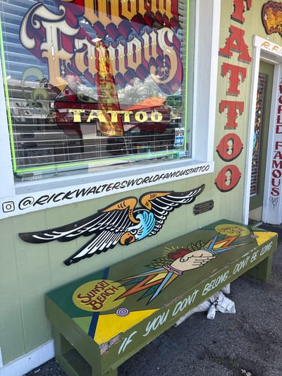 The image shows the exterior of Rick Walter's World Famous Tattoo shop in Huntington Beach, California. A red sign reading "TANNOUS" is partially visible above the shop's window, which has signs advertising that it is "OPEN" and inviting customers to "COME IN". A vertical sign to the side of the window reads "@RICKWALTERSWORLDFAMOUSTATTOO", next to a small Instagram logo. On the wall are decorations including a cutout of a tattoo design with wings and a painted sign for "SUNSET BEACH". The sign has the words "IF YOU DON'T BELONG DON'T BE LONG". A bench to the right is dedicated to Rick Walters with a note that says, "If you ain't gettin tattooed don't sit here".