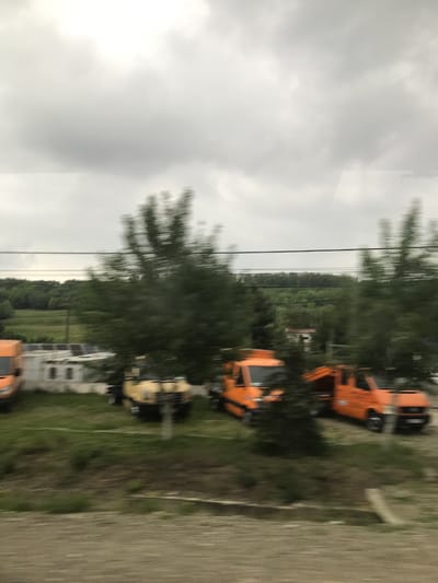 The image, taken in Cristești, Romania, presents an outdoor scene under a heavily overcast, light gray sky. Two dark, thin horizontal lines, consistent with utility wires, traverse the upper midground. The scene is characterized by significant motion blur, particularly affecting trees and ground elements, suggesting capture from a moving vantage point.
In the midground, a series of utility vehicles are parked on a grassy area. From left to right, a partial view of an orange van is visible. Immediately behind it and to its right stands a small, white, single-story building featuring multiple dark, rectangular window-like openings and a roof structure that appears to incorporate solar panels or a similar metallic installation. Further right, a yellow utility vehicle is partially obscured by vegetation. Adjacent to it are two distinct orange utility trucks, each equipped with specialized apparatus on its rear bed, possibly small cranes or lifting devices.
Numerous green trees, appearing blurred due to motion, are distributed across the midground and lower background. Beyond the parked vehicles and building, a flat green field extends, meeting a dense, dark green treeline or forest on the horizon. The immediate foreground consists of a blurred, textured surface of brownish-gray and green tones, consistent with an unpaved road or a grassy verge. No individuals are present in the frame.