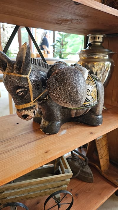 A close-up shot focuses on a dark, textured ceramic donkey figurine resting on a light-colored wooden shelf. The donkey is adorned with a yellow twine bridle and has a saddlebag on its back, decorated with a gold, black, and white patterned cloth. Behind the donkey, to the right, a shiny, silver samovar stands on the same shelf. Below this shelf, another wooden shelf holds a rustic wooden crate with black spoked wheels. Further back and to the right, resting on a lower shelf, are antique-looking cast-iron objects, possibly irons. In the background, through a window or opening, greenery and trees are visible, along with a blurred figure of a person wearing a dark shirt and headscarf. The scene appears to be set in a shop or a display area, possibly in the city of Krasnyy Kurgan, Russia.