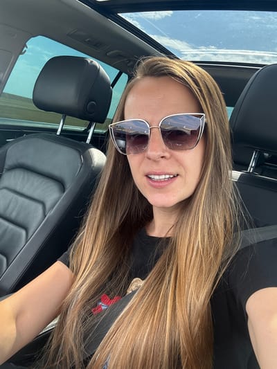 A woman with long, blonde-brown hair and sunglasses is sitting in the passenger seat of a car, with a sunroof open above her. She is wearing a black t-shirt with a red and blue embroidered design. The car's black leather interior is visible, including the headrest and seat. Outside the car, the landscape appears to be a flat, open field under a bright sky with some clouds. The image is taken from a slightly high angle, looking down at the woman. There is no indication of the city of Troyanovo, Bulgaria in the image.