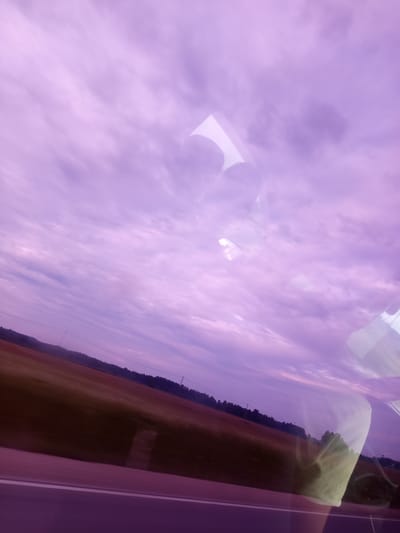 The image is a photograph taken from inside a vehicle, looking out through a window. The perspective is at an angle, with the bottom of the frame showing a road with a white line. Beyond the road, there is a field with a darker area, possibly a forest or trees, along the horizon. The sky is a prominent feature, filled with wispy, purple-toned clouds. There are reflections visible on the window surface, obscuring parts of the landscape and sky. These reflections appear to be of the interior of the vehicle and possibly a person or people. The overall lighting suggests it is either dawn or dusk, given the muted colors and cloud formations. There is no specific identifiable landmark or detail that places the image within the city of Salas, Latvia.