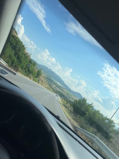 The image depicts a view from inside a vehicle traveling on a curved asphalt road. The road is bordered by a grassy embankment on the left and a guardrail on the right. Lush green trees and vegetation cover the left side of the road, extending up a hillside. In the distance, a valley unfolds with rolling hills, agricultural fields, and scattered buildings, suggesting a rural or semi-rural setting. The sky is a bright blue, adorned with scattered white cumulus clouds. The dashboard of the vehicle is partially visible in the foreground, with its instruments obscured by glare. The scene appears to be set in the municipality of Svetlina, Bulgaria, given the geographical context. There are no people or animals clearly visible in the image, and the action is the movement of the vehicle along the road. The overall environment is natural and scenic, characterized by a sunny day with good visibility.