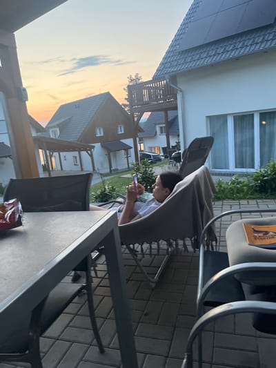 The image captures an outdoor patio scene at dusk, likely in the city of Milná, Czechia. In the foreground, a metal table with a dark surface is visible. To the right, a person is seated in a chair, wrapped in a brown blanket, and holding a smartphone. The person appears to be a young woman with dark hair, wearing a light-colored top. Behind her, a modern house with a white facade and a dark-tiled roof, some of which is covered with solar panels, is prominent. A wooden balcony or deck is attached to the house. The background shows more houses with similarly styled roofs, and a car is parked near one of them. The sky displays hues of orange and yellow, indicating sunset. The ground is paved with interlocking tiles.