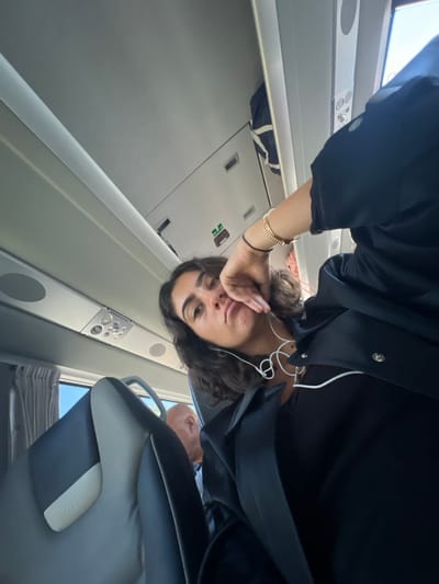 The image depicts the interior of a passenger transport vehicle, likely a bus or train, situated in the city of Pombal, Portugal. A young woman with dark, wavy hair is the primary subject, positioned at a low angle and looking directly at the camera. Her right hand rests against her cheek, and she wears a dark top, a dark jacket, a gold-beaded bracelet, and a thin black wristband. White wired headphones are draped around her neck. Behind her, partially visible to the left, is the bald head of another male passenger. The vehicle interior features upholstered seats in dark and light gray, light-colored overhead panels with air vents and reading lights, and a window on the left with drawn dark gray curtains, revealing bright daylight outside. An overhead storage compartment on the right contains a blue and white striped object.