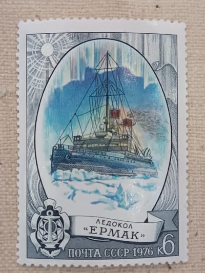 The image is a postage stamp from the USSR, dated 1976. The stamp features an illustration of an icebreaker ship, the "Ermak," navigating through icy waters under an aurora borealis. The ship is depicted centrally within an oval frame, with a sunburst design and stylized mountains in the background. At the bottom of the stamp, text reads "ПОЧТА СССР 1976 К" (Post of the USSR 1976 K) and "ЛЕДОКОЛ «ЕРМАК»" (Icebreaker "Ermak"). A circular emblem featuring an anchor and star is visible on the left side. The stamp is set against a textured, light brown background. There is no indication of the city of Onaldy, Turkmenistan in this image.