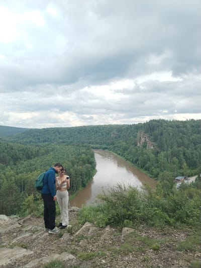 A man and a woman are standing on a rocky incline overlooking a wide, muddy river that curves through a densely forested valley. The man, wearing a blue jacket and black pants, has a green backpack slung over one shoulder. The woman, in a light-colored top and matching pants, is looking at a phone. They appear to be engaged in conversation. The sky is overcast with dramatic clouds. In the distance, a few buildings are visible near the riverbank, and the surrounding hills are covered in a thick carpet of green trees. The scene is likely set in the natural landscape near Idrisovo, Russia, known for its picturesque river valleys and forests.
