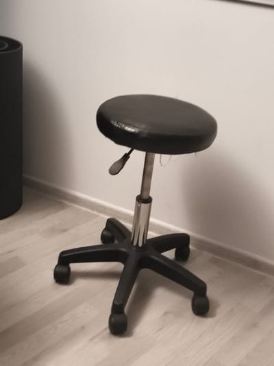 A black, round-topped stool with a metal adjustable height mechanism and a black five-legged base with casters sits on a light-colored wooden floor. The stool is positioned near a plain, light-colored wall with a white baseboard running along the floor. To the left of the stool, a dark gray cylindrical object, possibly a trash can or a planter, is partially visible. The scene is indoors, in a room with neutral-colored walls and flooring. There is no indication of the specific location within Troisdorf, Germany.