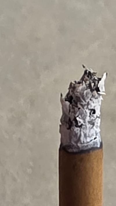 The image is a close-up, head-on shot of the burning end of a cigarette. The ash is a pale gray and appears somewhat fluffy, with some darker charred bits mixed in. The cigarette paper is visible as a thin white band just below the ash. The body of the cigarette is a light brown color, consistent with tobacco. The background is out of focus and appears to be a textured, neutral-colored wall. There are no people or specific environmental details visible that would allow for the identification of a location in Novyy Chirkey, Russia.