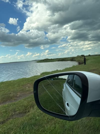 The image depicts a natural outdoor scene, identified as being in Айдарлы, Kazakhstan. The perspective is from or immediately adjacent to the passenger side of a white vehicle, with a prominent view of its side mirror in the lower right foreground.
The environment features a large body of water occupying the mid-left portion of the frame, bordered by a green, grassy bank in the foreground and midground. The water exhibits surface ripples, indicative of wind. The sky above is partially cloudy, showing areas of blue sky interspersed with both bright white and dark grey cumulus and stratocumulus clouds.
On the grassy bank to the right, a solitary individual, dressed in dark attire, stands facing the water's edge, partially obscured by the vehicle's side mirror. Two red fishing rods are visible angled towards the water from the grassy bank, positioned to the left of the individual.
The side mirror reflects the white body of the vehicle, including a door handle and side window, as well as the grassy terrain behind the vehicle where a small, light-colored tent is discernible, suggesting a camping setup. The ground in the immediate foreground, outside the vehicle, consists of green grass with visible worn paths or dirt patches. In the distance across the water, the landscape features low-lying vegetation or brush under the cloudy sky.