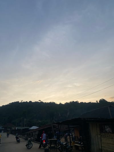 The image captures a roadside scene in Igarra, Nigeria, at either dawn or dusk. The sky transitions from a light blue-grey at the top to a pale golden-yellow near the horizon, exhibiting scattered, thin cloud formations. A large, densely forested hill or mountain, appearing in dark silhouette, dominates the mid-background.

In the lower half of the image, an unpaved road is visible, on which several individuals and motorcycles are present. On the right side of the road, multiple rudimentary structures with dark, corrugated iron roofs are prominent, largely obscured by shadow. Power lines span diagonally across the upper right portion of the image.

Specific subjects include approximately seven motorcycles, some parked and others in motion, and at least eight discernible individuals. One person wearing a red hooded garment and dark pants is seated on a motorcycle, facing away from the viewer, positioned prominently on the right side of the road. Another individual in yellow attire is visible near one of the structures on the right. Other people are standing or walking along the road and near the buildings. The overall lighting conditions are low, producing deep shadows and emphasizing silhouettes of the people, vehicles, and structures against the brighter sky.