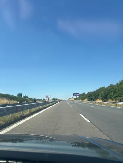 This image shows a view from a vehicle driving on a highway under a clear blue sky. The highway has multiple lanes and is bordered by a metal guardrail and vegetation. There are two billboards visible in the distance along the highway. One billboard on the left side of the road has the text "imot.bg" on it. The other billboard on the right side has the text "МЕБЕЛИ Carmen". The scene appears to be in a rural or suburban area, possibly on the outskirts of the city of Vratitsa, Bulgaria, given the presence of the billboards which are likely advertisements for local businesses. The camera is positioned low, suggesting it is mounted on the dashboard of a car.