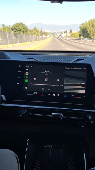 The image displays a daytime view from inside a vehicle traveling on a multi-lane highway, identified as being in the vicinity of San Rafael Tlanalapan, Mexico.
In the foreground, the interior of a vehicle is visible, focusing on a large digital infotainment screen. The screen displays a music application showing the track "Duro" by "Skrillex, Young Miko" currently playing, with 2 minutes and 37 seconds elapsed and 29 seconds remaining. The time "8:53" and "5G" cellular connectivity are visible in the upper left corner of the screen. Navigation, telephone, and media options are present on the left side of the display. Below the screen, climate control elements are visible, including "OFF" indicators. A portion of a person's leg, clad in light-colored pants, is visible in the lower-left corner of the frame.
The midground features a straight, paved highway with multiple lanes, marked by yellow and white lines, extending towards the horizon. A concrete barrier with yellow markers separates the opposing directions of traffic (not fully visible) or the highway's edge. A black mesh fence runs parallel to the highway on the left side, backed by dense green trees and vegetation. Several vehicles, including a motorcycle, are visible on the highway in the distance.
The background reveals a clear, light blue sky above a range of distant mountains, some exhibiting lighter coloration on their peaks, possibly indicative of snow or rock. The overall environment suggests clear weather conditions.