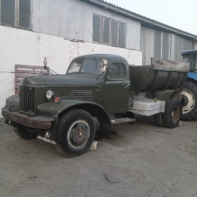 This image depicts an outdoor scene with a vintage military-style truck parked in front of a weathered building. The truck is dark green and appears to be a utility vehicle, possibly a dump truck, with a large cargo bed in the rear. It has a distinct grille, round headlights, and large tires. To the right of the truck, a portion of a blue tractor is visible, suggesting an agricultural or industrial setting.
The building behind the truck is constructed of light-colored material, possibly stucco or painted concrete, with corrugated metal siding further down. There are several windows with wooden frames on the building, some of which appear to be reflective or covered. The ground is a rough, gravelly surface, and the overall impression is one of a utilitarian or storage area.
There is no discernible activity taking place in the image; the vehicles and building appear stationary. Notable details include the age and condition of the truck, which shows signs of wear and tear, and the weathered appearance of the building. The lighting suggests it is either early morning or late afternoon, with a diffused, even light. The sky is overcast. There is no visible text in the image. The location is Ploske, Ukraine.