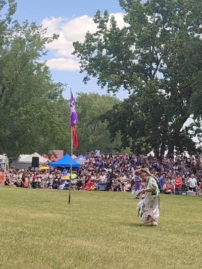 The image shows an outdoor gathering in Kahnawake, Canada, with a large crowd of spectators seated on bleachers and on the grass. In the foreground, a person in traditional Indigenous regalia, adorned with feathers and intricate beadwork, is performing a dance. The person is wearing a long, flowing skirt and a decorated top. In the background, two flags are visible on a pole, one purple with a white emblem and another red. Tents and structures suggest a festival or event setting. Lush green trees frame the scene under a partly cloudy blue sky.