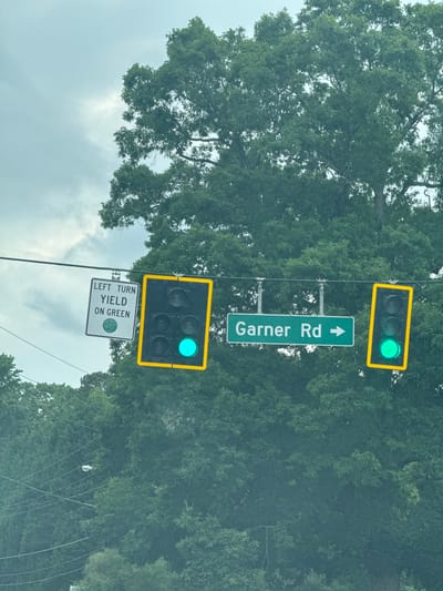 The image depicts a street intersection in Mountain Park, United States of America. A street sign reading "Garner Rd" with an upward-pointing arrow is visible. Above the sign is a traffic light displaying a green light. Below the sign is another traffic light also displaying a green light, and a sign that reads "LEFT TURN YIELD ON GREEN". The background consists of trees and a cloudy sky.
