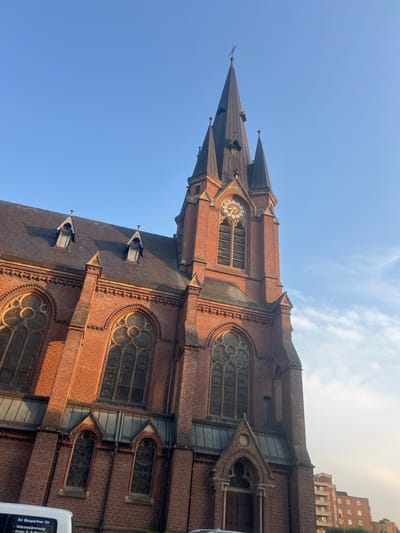 This is a vertically oriented, low-angle shot of a large brick church with a tall, pointed spire and a clock face. The church is made of red brick and features numerous arched windows, some with decorative tracery. The spire has several smaller spires or finials. The sky above is a clear blue with a few wispy white clouds, suggesting fair weather. The sun appears to be low in the sky, casting long shadows on the roof and illuminating the brickwork with a warm glow, indicating either early morning or late afternoon. The building's architecture suggests a Gothic or Neo-Gothic style. In the background, to the right of the church, parts of a modern, multi-story building are visible.
There are no people visible in the image. The primary object is the church building. The scene is an exterior view of the church under a clear sky. The overall impression is one of grandeur and historical significance.
A small amount of text is visible on the white vehicle in the lower right corner of the image, which appears to be a van. The text is in German and reads: "Ihr Baupartner für - Wärme Dämmung - Innen & Außen". This translates to "Your construction partner for - thermal insulation - interior & exterior".
The provided location context states Herne, Germany, which, combined with the German text, suggests the church is located in Herne, Germany.