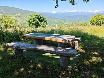 An outdoor wooden picnic table and bench set in a grassy area with a mountainous landscape in the background. The table and bench are made of rough-hewn logs. The landscape features rolling green hills, dense vegetation, and snow-capped mountains in the distance under a clear blue sky. The scene appears to be in a rural or mountainous region. It is possible this location is near Ukeshi, Georgia, known for its scenic mountain vistas.