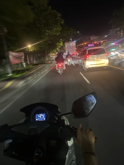 An out-of-focus, first-person perspective night-time shot captures a person riding a motorcycle in heavy traffic on a street in Denpasar Barat, Indonesia. The motorcycle's digital speedometer displays a speed of 47 km/h. Ahead, a white car is visible with its brake lights illuminated, along with multiple other vehicles, including a truck and several motorcycles. Trees line the right side of the road, and streetlights cast a warm glow on the scene. The overall impression is one of a bustling urban environment at night.