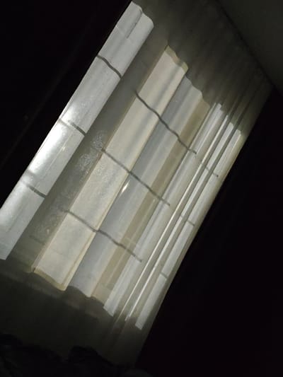 The image is a high-angle, dimly lit view of a slatted surface, possibly a translucent roof or ceiling, with light filtering through in vertical strips. Dark shadows dominate the edges and upper portions of the frame. The slats appear to be made of a corrugated material, likely plastic or fiberglass, with some speckling or texture visible on their surface. There are darker horizontal lines crisscrossing the slats at intervals. The overall impression is one of diffused light entering an enclosed or partially enclosed space. The image does not contain any elements that would allow for an identification of its location in Chernysevka, Russia, nor does it depict any people, actions, or interactions. It is a still, environmental shot.