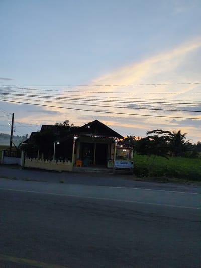 The photograph shows a roadside scene in Teupok Tunong, Indonesia, taken at dusk or dawn. A one-story building stands to the side of the road, likely a small business, indicated by the sign visible to the right of the entrance. The building has a dark roof, possibly made of tile or thatch, with a porch area illuminated by a string of lights. In front of the building are at least two chairs, one orange and one green. A low white fence partially surrounds the building.

The sky displays a blend of blue and orange hues, suggesting either sunrise or sunset. Several power lines stretch across the upper portion of the frame. To the right of the building, there is vegetation. The road in the foreground occupies a significant portion of the image. In the background, trees and what appears to be a field can be seen. The lighting suggests a low level of natural light augmented by artificial illumination from the building. No people are visible in the photograph.