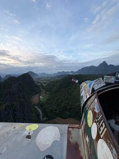 An overhead shot captures a breathtaking vista of lush green mountains and valleys in Ban Phon Sai, Laos. The foreground is dominated by the rusty, worn exterior of a hot air balloon basket, adorned with a collage of colorful stickers. Among them, a yellow oval sticker reads "Nett hier, aber waren sie schon im Narakeller?" which translates to "Nice here, but have you been to the Narakeller?". The landscape unfolds with rolling hills, a winding road, and patches of farmland. In the distance, several hot air balloons drift lazily across the pale blue sky, adding a touch of whimsical adventure to the serene natural beauty. The clouds are wispy and scattered, hinting at the early morning light.
