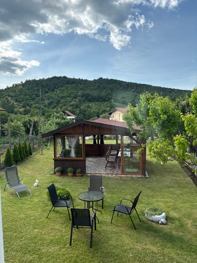 The image shows a lush green backyard with a wooden gazebo attached to a house. A table with four chairs is set on the grass. A ceramic stork statue sits in the grass nearby. In the background, a forested hill rises, and a partly cloudy sky is visible. The location is Polena, Bulgaria.
