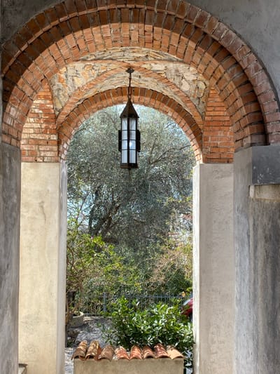 The image displays an architectural passage in Vrijesno, Montenegro, framed by a prominent brick archway. The arch is constructed from reddish-brown bricks, with some exposed rough stone elements integrated into its structure, and is supported by plastered columns; the left column is light-colored, and the right is darker and exhibits weathering. A black, metal-framed lantern, featuring four frosted glass panes, hangs centrally from the arch's apex and is illuminated from within. Below the lantern, a low concrete wall is topped with a row of approximately seven reddish-orange terracotta roof tiles. Through the arch, an outdoor garden scene is visible. A large tree with silvery-green foliage, consistent with an olive tree, occupies the background. Below this tree, dense green shrubs and smaller plants are present. A dark metal fence is partially discernible behind the lower vegetation, and a small section of a red object is visible beyond the fence on the right. The scene is illuminated by natural light, and no individuals or specific actions are depicted.