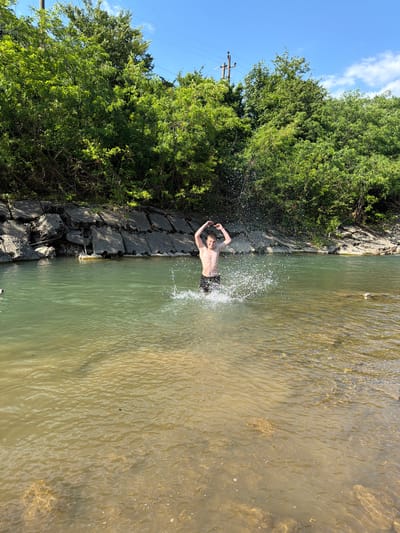 A shirtless young male is standing in shallow, clear water, splashing enthusiastically. He is wearing dark swim trunks and has his arms raised in a "V" shape. Water droplets are airborne around him, indicating active splashing. The background features a natural setting with lush green trees lining the bank and a clear blue sky overhead. The water is a mix of green and sandy-colored depths. The location is identified as the city of Krasnoye, Russia.