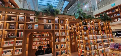 The image depicts the interior of a large, modern bookstore in Gwangmyeong, South Korea. The bookstore features extensive wooden shelving units filled with numerous books. The shelves are illuminated by warm, embedded lighting, creating an inviting atmosphere. Some shelves are dedicated to specific genres or themes, indicated by signs such as "세계 문학전" (World Literature) and "학술지" (Academic Journal).
In the foreground, two individuals are seated facing a bookshelf within an alcove-like seating area. They appear to be browsing or reading, with one person holding a device. The overall environment is well-lit, with natural light filtering in from a large skylight or ceiling structure visible at the top of the image. Lush green plants are strategically placed on top of some shelving units, adding a touch of nature to the indoor space. The bookstore is multi-leveled, with visible railings and balconies suggesting upper floors. The display of books suggests a wide selection catering to various interests.