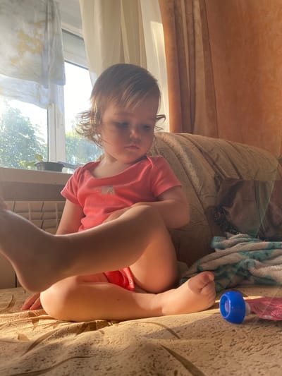 The image shows a young child sitting on a couch. The child is wearing a pink t-shirt and has blonde hair. The child is looking at the camera with a serious expression. The couch is upholstered in a beige fabric with a textured pattern. Behind the child, there are curtains and a window. Sunlight is streaming through the window, indicating it is daytime. A blue object, possibly a bottle cap or toy, is visible on the couch to the right of the child. There is also a folded blanket with a blue and white pattern on the couch. The overall setting appears to be a living room.
