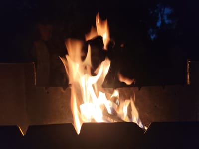 The image depicts a campfire at night in Kolpakovo, Russia. The primary subject is the fire itself, with vibrant orange and yellow flames illuminating the scene. The fire is contained within a metal fire pit, with dark, indistinct shapes suggesting the surrounding environment. The background is predominantly dark, indicating nighttime. The flames are dynamic and energetic, suggesting a lively fire. There are no discernible people in the image, and no text is visible. The setting appears to be outdoors, possibly in a rural or natural area, given the darkness and lack of urban features. The overall mood is warm and inviting, despite the darkness.