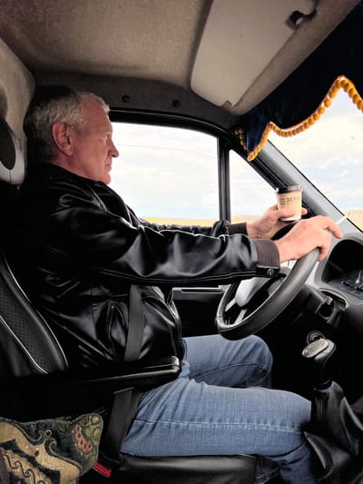 The image captures a scene inside a vehicle, likely a truck cab, with a man resting or lying down. The main subject is an older man with gray hair and a short beard, wearing a black leather jacket. He is positioned on his back, seemingly across the seats or a bunk area, with his head towards the rear of the cabin and his body extending towards the front. His hands are raised near the steering wheel, holding a disposable cup. Another person's legs, clad in blue jeans, are visible to the right, suggesting they are seated in an adjacent area.
The setting is a vehicle interior, characterized by beige paneling on the ceiling and walls, and a steering wheel. A dark blue decorative curtain with yellow pom-poms hangs near the windshield, indicating a personalized or long-haul vehicle. Through the windshield, a bright, overcast sky is visible, suggesting daytime.
The man appears to be at rest, possibly taking a break from driving. The cup he is holding has the Russian word "ЗЕРНО" (meaning "GRAIN") printed on it, along with "000". A black travel bag or duffel bag is seen on a seat beside him. A patterned cushion or blanket in shades of green, gold, and red is also visible near the feet of the person in jeans. The overall atmosphere is one of calm and respite inside a travel environment in Meshcheryaki, Russia.