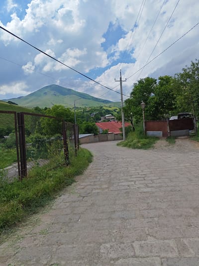 The image depicts a cobblestone road leading uphill in the city of Bazum, Armenia. The road is lined with a rusty metal fence on the left and tall trees and a gate on the right. In the distance, a lush green mountain looms under a cloudy sky. Several houses with red roofs are visible in the valley. Overhead, power lines crisscross the sky. The scene appears tranquil and rural.