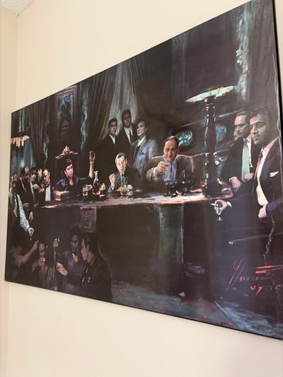 The image depicts a reproduction of a painting hung on a light-colored wall. The painting shows a crowded scene inside a dimly lit room. Numerous men are seated or standing around a long table. Some men are drinking, smoking, and interacting. There are bottles and glasses on the table. A portrait hangs on the wall behind some of the men. The style suggests a painting that depicts an event or scene from a historical or fictional depiction of a mafia party, possibly related to The Godfather movies. The location mentioned, Woodstock, Canada, has no significance or relationship to the artwork, which depicts an interior setting.