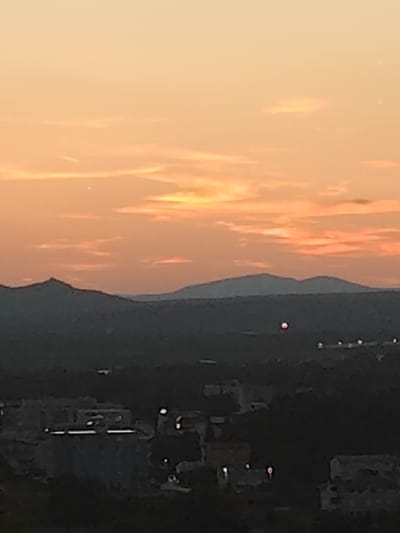 This is a long-range, low-resolution photograph of a landscape at dusk, likely taken from an elevated position overlooking a town. The sky is a gradient of warm orange and yellow hues, with thin wispy clouds scattered across it. In the mid-ground, silhouetted mountains form a dark, jagged horizon line. Below the mountains, the landscape transitions into a darker, indistinct area that appears to be a town or city. Numerous small, bright lights are visible in this area, suggesting artificial illumination from buildings or streetlights. In the center of the image, a small, bright red circular object is visible in the sky, and several other smaller, white lights are scattered throughout the darker landscape. The image quality is poor, with significant noise and blur, making detailed identification of specific structures or objects challenging. The scene depicts a rural or semi-rural setting at twilight, with a prominent natural landscape element (mountains) dominating the background and a populated area in the foreground.