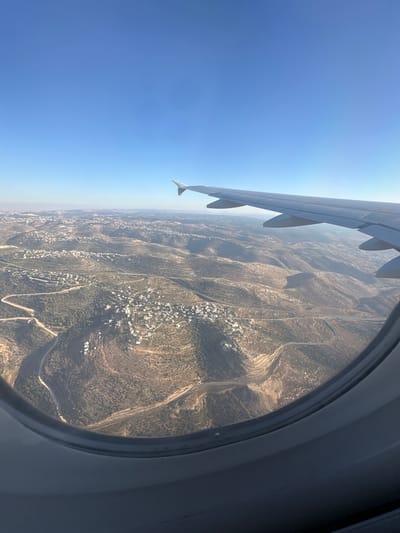 The image is an aerial view captured from an airplane window, looking down at a landscape that appears to be mountainous and arid. A portion of the airplane's wing is visible in the upper left of the frame, extending diagonally across the image. The sky is a clear, bright blue, indicating it's daytime.

Through the circular airplane window, the ground below is textured with rolling hills and valleys. Sparse vegetation, appearing as scrub and trees, dots the terrain. There are settlements with clusters of white buildings nestled within the landscape, suggesting human habitation. Roads are visible winding through the hills, connecting these settlements. The overall impression is one of a rural, perhaps semi-arid or Mediterranean environment.

The time of day is likely morning or afternoon due to the bright sunlight and clear skies. There are no visible people or specific objects on the ground that stand out, beyond the general features of the landscape and the settlements. The perspective is from a significant altitude, as is typical for aerial photography from a commercial aircraft. No text is visible in the image.