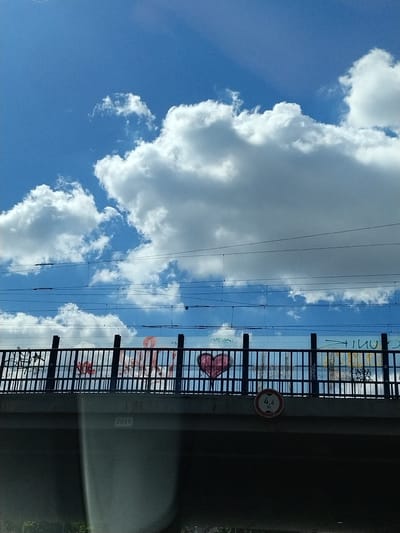 A low-angle, exterior shot captures a bridge in Strakonice, Czechia, under a bright blue sky filled with fluffy white clouds. Power lines are strung across the sky above the bridge. The bridge itself is constructed from concrete and features a black metal railing along its top. Graffiti adorns the side of the bridge, including a pink heart and various text markings. Below the railing, a circular sign indicates a height restriction of 4.6 meters. The year "2015" is visible on the concrete support structure of the bridge. The scene appears to be captured from street level, looking upwards at the bridge structure and the sky.