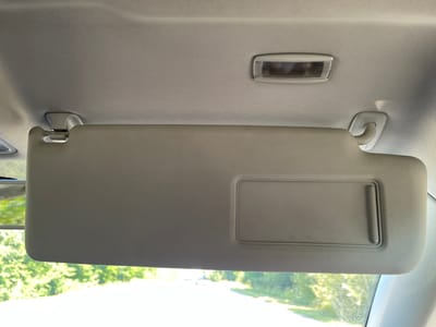 The image shows the interior of a car, specifically focusing on the passenger-side sun visor. The sun visor is a light gray color and has a rectangular vanity mirror inset into its surface. It is attached to the car's headliner by two white plastic brackets, one on the left and one on the right. Above the sun visor, on the headliner, is a rectangular interior light fixture with a clear lens. The headliner itself has a textured, woven fabric pattern. Through the car's windshield, which is visible below the sun visor, is a blurred view of green trees and a road, suggesting an outdoor environment, likely during the daytime. The provided location of Maklaivka, Ukraine, is not visually represented in the image itself.