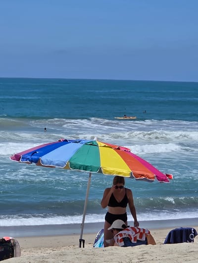 The image depicts a sunny day at the beach, likely in San Clemente, California, given the characteristic surf and sand. The foreground features a colorful, multi-panel beach umbrella providing shade. Beneath the umbrella, two individuals are present. A woman wearing a black bikini and sunglasses stands, adjusting her sunglasses. Next to her, a person wearing a white cap and a towel draped over their shoulders is positioned lower down on the sand. In the background, the ocean displays moderate waves and the horizon shows a clear blue sky. Several surfers can be seen in the water, with one person lying on a surfboard. The overall scene conveys a typical beach day with people enjoying recreational activities.
