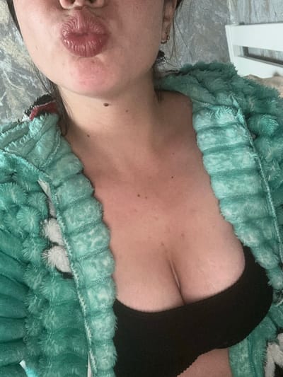 The image is a close-up, over-the-shoulder shot of a person wearing a plush, teal robe with white accents, and a black bra. The person's face is partially visible, with their lips puckered in a kiss. Their chest is also visible, with cleavage showing. The background is blurred, but appears to be an indoor setting with a bed and a white piece of furniture, possibly a dresser or headboard. There is no discernible connection to Gavrilov Posad, Russia.