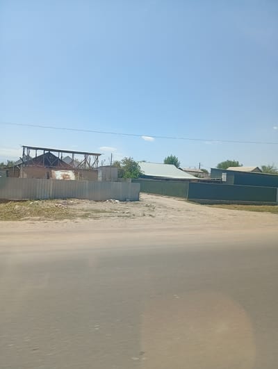 The image depicts a rural scene in the village of Екпінді, Kazakhstan. In the foreground, a dusty road is visible, suggesting a dry, possibly arid environment. Beyond the road, there are several structures, including a house under construction with exposed wooden beams and brick walls, enclosed by a corrugated metal fence. Further in the background, other buildings with corrugated metal roofs and fences are present, some appearing to be warehouses or industrial sheds, suggesting a mix of residential and possibly commercial or agricultural use. The sky is clear and blue with a few wispy clouds, indicating a sunny day. Trees are scattered throughout the landscape, adding to the rural character of the scene. There are no people or vehicles clearly visible in the image. The overall impression is one of a developing or established rural community in Kazakhstan.