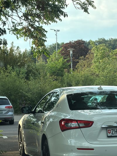The image shows a close-up view of the rear of a white car, likely a Maserati, parked outdoors. The car is in the foreground, with its taillight and license plate visible. The license plate has "ZH" followed by a Swiss flag and numbers. The car is parked on a paved surface, possibly a street or parking lot, adjacent to a wooded area.

Behind the car, the scene transitions to a lush green environment with various trees and foliage. Sunlight filters through the leaves, creating dappled patterns on the car's surface and suggesting it is daytime. The sky is a pale blue with some wispy clouds. In the distance, a tall pole with an antenna-like structure is visible among the trees, possibly indicating a communication tower or street light. The overall atmosphere is peaceful and natural. The image is oriented vertically, which is unusual for a typical photograph of this nature, suggesting it might have been taken from a phone held in portrait mode and later rotated. There are no people visible in the frame.