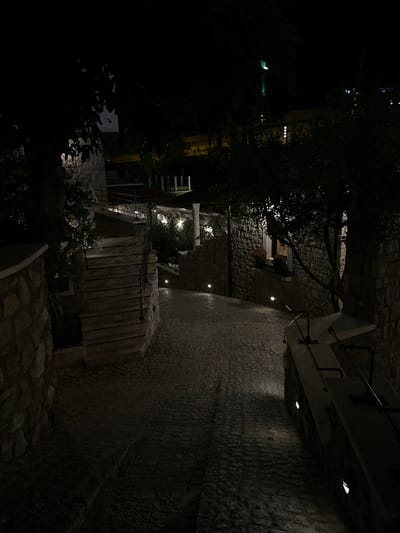 The image depicts a nighttime view of a cobblestone path and stone staircase in Bartula, Montenegro. The path is illuminated by several small, downward-facing lights embedded in the stone walls on either side. A large stone wall runs along the left side of the path, and a stone staircase with metal railings is visible on the left, ascending away from the viewer. On the right side, another stone wall is visible, with a tree growing beside it. In the background, glimpses of buildings and distant lights suggest an urban or semi-urban environment. The overall scene conveys a sense of quiet and ambiance due to the low lighting and the textured stone architecture.